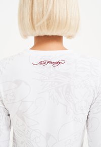 Valkoinen pitkähihaisshirt, jossa on vaaleanharmaita tiikeri- ja tekstikuvioita, sekä punainen "Ed Hardy" -logo selässä lähellä kaula-aukkoa.