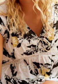 Vestido blanco con estampado floral negro y acentos amarillos. Con un escote en V, detalles de encaje y un lazo en el escote. Tejido texturizado.