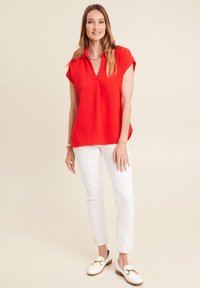 Top rosso a maniche corte con scollatura a V e tessuto testurizzato, abbinato a pantaloni bianchi slim-fit e mocassini bianchi con un accento dorato.