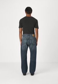 Camiseta de color gris oscuro combinada con jeans de denim azul de corte holgado que presentan un parche de cuero y bolsillos traseros detallados, vistos desde la parte trasera.