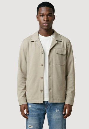 Mężczyzna w beżowej kurtce zapinanej na guziki, na białej koszuli i podartych niebieskich dżinsach, stojący na tle jasnego, jednolitego tła.