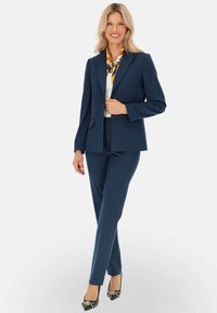 Granatowy garnitur z dopasowaną marynarką i obcisłymi spodniami. Biała bluzka i kolorowy szal podkreślają całość. Czarne szpilki z dekoracyjnym wykończeniem.
