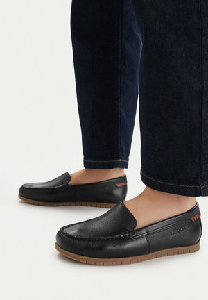 Mocassini neri in pelle senza lacci con suole in gomma marrone, indossati con jeans blu scuro risvoltati e piedi nudi.