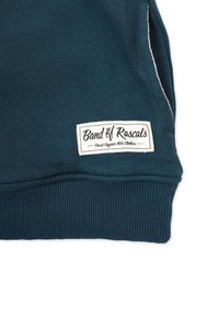 Tumma turkoosi hupparin kangas, jossa on ribattu hihansuukappale. Valkoinen etiketti lukee "Band of Rascals" ja "Finest Organic Kids Clothes", kiinnitetty helmassa.