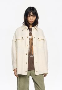 Joven vestido con una chaqueta de botones crema de gran tamaño, una camiseta gráfica marrón y pantalones holgados verde oliva, de pie frente a un fondo blanco.