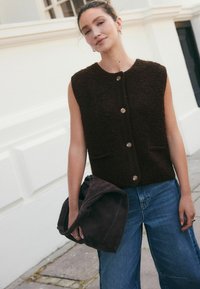 Bruine, mouwloze vest met textuurstof en frontzakken, met een gouden knoopsluiting, gecombineerd met blauwe wijde jeans en een donkere tas.