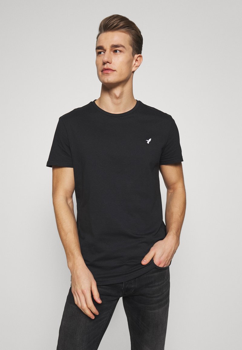 Camiseta negra de manga corta hecha de algodón, con cuello redondo y un pequeño logotipo blanco en el lado izquierdo del pecho. Combinada con jeans oscuros.