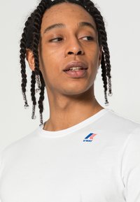 Camiseta blanca de manga corta con el logo de K-Way en azul y rojo en el hombro izquierdo, hecha de una tela suave y transpirable.