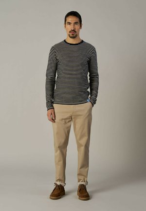 Langarmshirt in Marineblau und Creme mit Streifen, kombiniert mit beigen Hosen und braunen Wildlederschuhen; hat ein blaues Etikett auf dem Shirt.