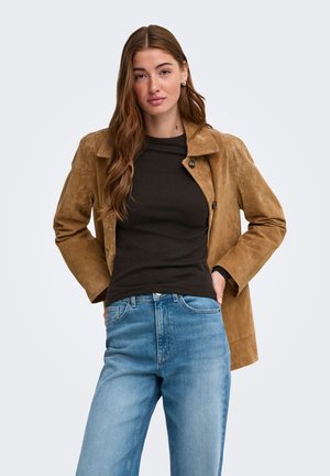 Ung kvinde med langt brunt hår iført en tan farvet ruskindjakke, sort skjorte og blå jeans, stående med hænderne i lommerne.
