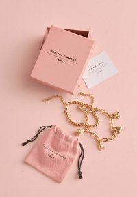 Guld charmerarmbånd med forskellige former, præsenteret i en lyserød æske. Inkluderer en posetaske med snorelukning og et 'Tak' kort.