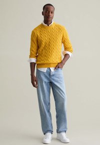 Gelber Kabelstrickpullover über einem weißen Hemd, kombiniert mit hellblauen Jeans und weißen Sneakers. Der Pullover hat eine strukturierte Verarbeitung.
