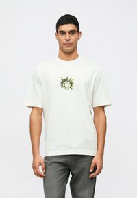 Ung man som bär en vit T-shirt med en svart och gul solansiktsgrafik, tillsammans med urtvättade svarta jeans, stående framför en enfärgad bakgrund.