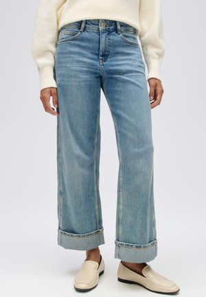 Person trägt hellblaue, weit geschnittene hochgekrempelte Jeans, cremefarbene Slipper und einen cremefarbenen Pullover mit langen Ärmeln, steht vor einem einfarbigen Hintergrund.