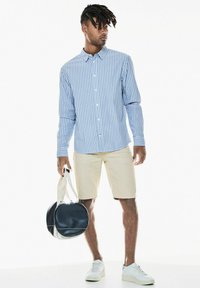 Světle modrá a bílá pruhovaná košile na knoflíky, béžové šortky, bílé tenisky a černo-bílá kulatá taška.