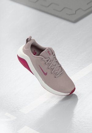 Licht mauve Nike hardloopschoen met roze swoosh en accenten op de zool zwevend boven een grijs oppervlak met witte markeringen.
