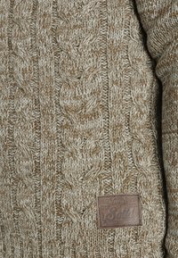 Beige-Kabelstrickpullover mit strukturierten Mustern, einem gerippten Kragen und einem Lederpatch mit der Aufschrift "Solid" auf der Vorderseite.