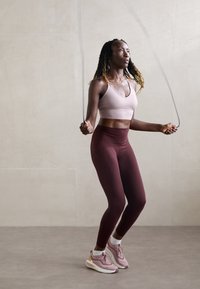 Atleta in un reggiseno sportivo rosa chiaro e leggings bordeaux, che salta con una corda. Indossa scarpe da ginnastica rosa chiaro e bianche.