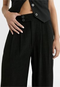 Zwarte wijde broek met plooien, met een hoge taille en twee zwarte knoopsluitingen. Het materiaal heeft een licht gestructureerde afwerking.