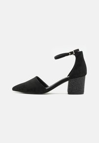 Tacones altos de ante negro con tiras en el tobillo, punta afilada y tacón grueso texturizado. Presenta un interior suave y una correa ajustable.