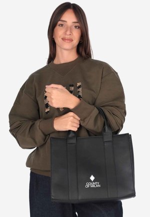 Donna con lunghi capelli scuri che indossa un maglione marrone e tiene una borsa nera "County of Milan" con doppi manici.