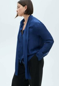 Blusa di seta blu navy con vestibilità ampia, maniche lunghe e un nodo drappeggiato al collo. Presenta polsini con bottoni e una texture liscia.
