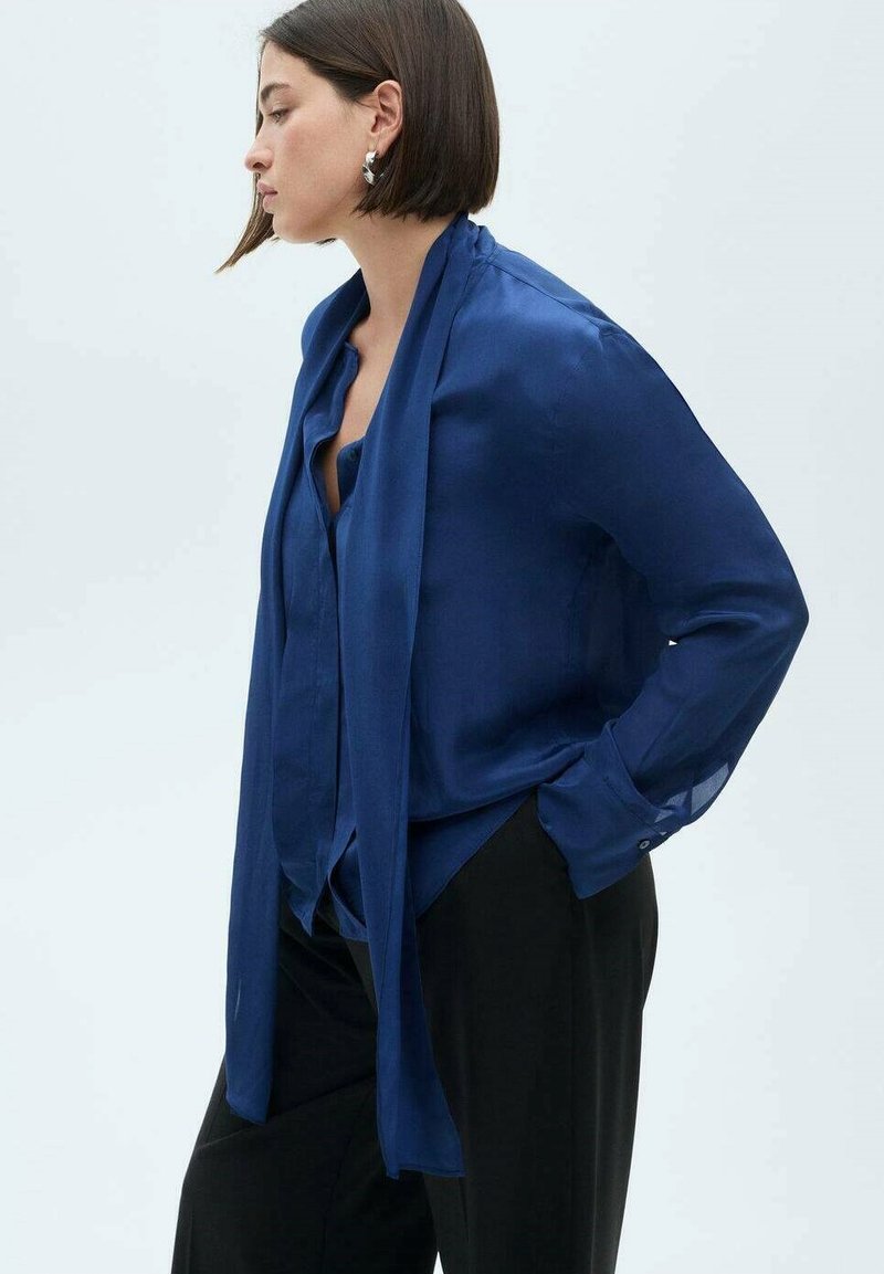 Blusa di seta blu navy con vestibilità ampia, maniche lunghe e un nodo drappeggiato al collo. Presenta polsini con bottoni e una texture liscia.