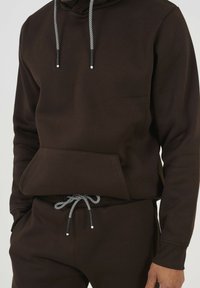 Ciemnobrązowa bluza z kapturem z kieszenią z przodu, kontrastującymi czarnymi i białymi sznureczkami oraz luźnym krojem, w połączeniu z pasującymi ciemnobrązowymi spodniami.
