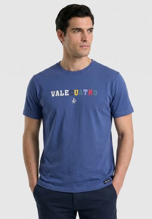 Hombre con una camiseta azul de manga corta con "VALECUATRO" en letras multicolores, de pie con las manos en los bolsillos y mirando a la derecha.