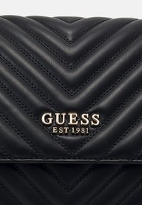 Czarna skórzana torba z pikowanym wzorem w jodełkę, z nazwą marki "GUESS" i napisem "EST 1981" w złotych metalowych ozdobach.