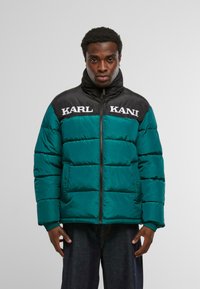 Pikowana kurtka w kolorze zielonkawoniebieskim z czarnym karczkiem na ramionach, zapinana na zamek z przodu, z białym napisem "KARL KANI" na piersi. Ściągacze na mankietach i pikowana faktura.