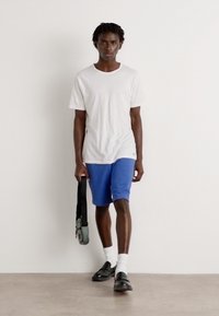 Tricou alb cu mânecă scurtă, pantaloni scurți albăstrii, încălțări negre și geantă neagră. Modelul poartă șosete albe; fundalul este neutru și minimal.
