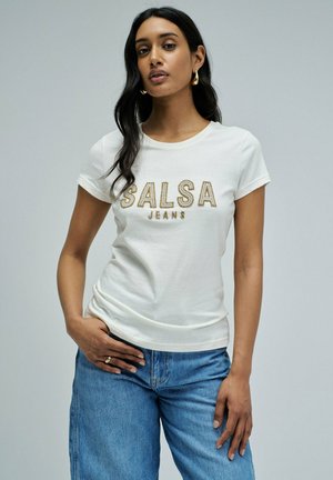 Mujer con cabello largo y oscuro, que lleva pendientes de aro dorados, una camiseta blanca de "SALSA JEANS" y jeans azules, posando con la mano en la cadera sobre un fondo gris.