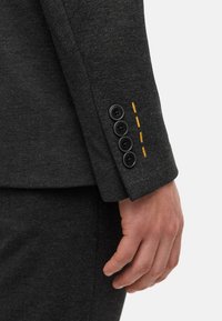 Mørk grå skreddersydd dressjakke med ermer som har fire sorte knapper. Gule stingdetaljer nær knappene gir en ekstra touch. Mykt teksturert stoff.