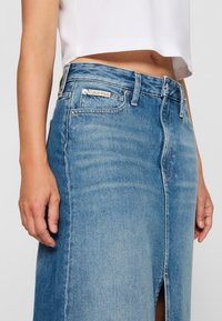 Šviesiai mėlyna džinsinė sijonas su priekinėmis plyšiu, klasikinis penkių kišenėlių dizainas ir Calvin Klein etiketė ant liemens.