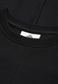 Un primo piano del colletto di una maglietta nera, con colletto a coste e un'etichetta bianca con un logo sottostante.