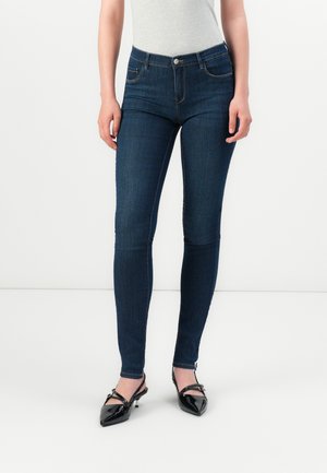 Woman wearing dark blue skinny jeans and black pointed-toe heels standing against a plain white background.