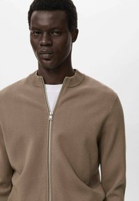 Man met kort gekruld haar in een lichtbruine rits trui over een wit shirt, staande tegen een effen lichtgrijze achtergrond.