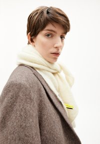 Bruine jas met structuur en een crème sjaal om de hals gewikkeld. De sjaal heeft een zachte, fluweelachtige textuur en een zichtbaar geel labeldetail.