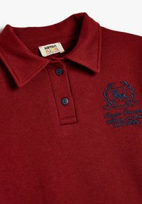 Bordowa bawełniana koszulka polo z kołnierzykiem, wyposażona w dwa granatowe guziki oraz haftowany wzór lwa w dekoracyjnej laurowej gałązce.