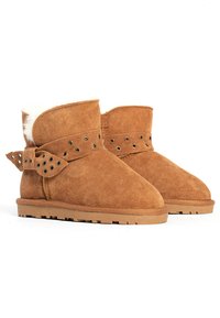 Botines de ante color marrón claro con un suave forro de piel de oveja blanca. Presentan una correa decorativa con recortes circulares y un lazo en la parte de atrás. Suela plana.