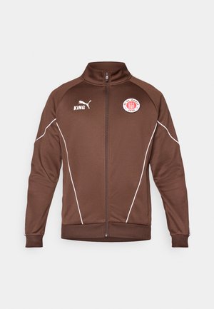 Braune Zip-Jacke aus glattem Material. Sie verfügt über weiße Akzentlinien, einen Stehkragen und Logos für KING und FC St. Pauli.