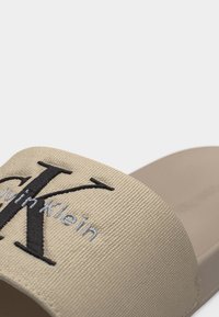 Μπεζ σαγιονάρα με υφασμένη λουρίδα που διαθέτει μεγάλο μαύρο γράμμα "K" και μικρότερο γκρι κέντημα με το λογότυπο "Calvin Klein".