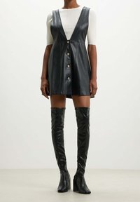 Vestido sin mangas de cuero negro con escote en V, con una fila de botones plateados. Combinado con botas negras hasta la rodilla, de textura suave.