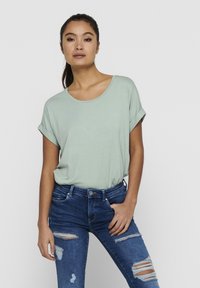 Helles grünes T-Shirt mit kurzen Ärmeln und rundem Ausschnitt. Kombiniert mit engen blauen Jeans mit Rissen und mittelhoher Taille.
