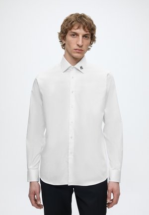 Junger Mann mit lockigem Haar, der ein weißes Hemd mit Knöpfen und einem kleinen schwarzen Logo am Kragen trägt, steht vor einem schlichten hellen Hintergrund.