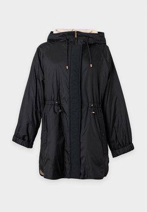 Femme portant un parka noir à capuche avec cordons, un pantalon beige à jambes larges et des bottines noires, debout devant un fond uni.