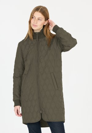 Femme portant une longue veste matelassée vert olive avec des poignets côtelés, debout, une main touchant ses cheveux.