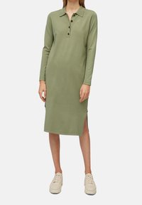 Robe verte à manches longues avec un col, patte de boutonnage, fentes latérales et texture douce, assortie à des chaussures de couleur claire.