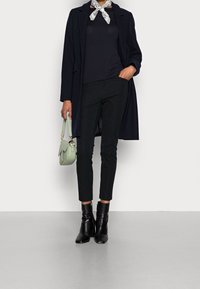 Schwarzer Mantel über einem marineblauen Oberteil, schwarze schmale Hosen und schwarze Knöchelstiefel. Eine hellgrüne Handtasche mit einem kurzen Griff und ein gemustertes Tuch runden den Look ab.